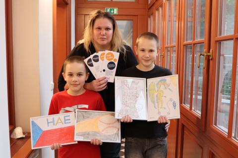 Na fotografii jsou dva chlapci a jedna žena, ukazují obrázky a letáčky organizace HAE Junior